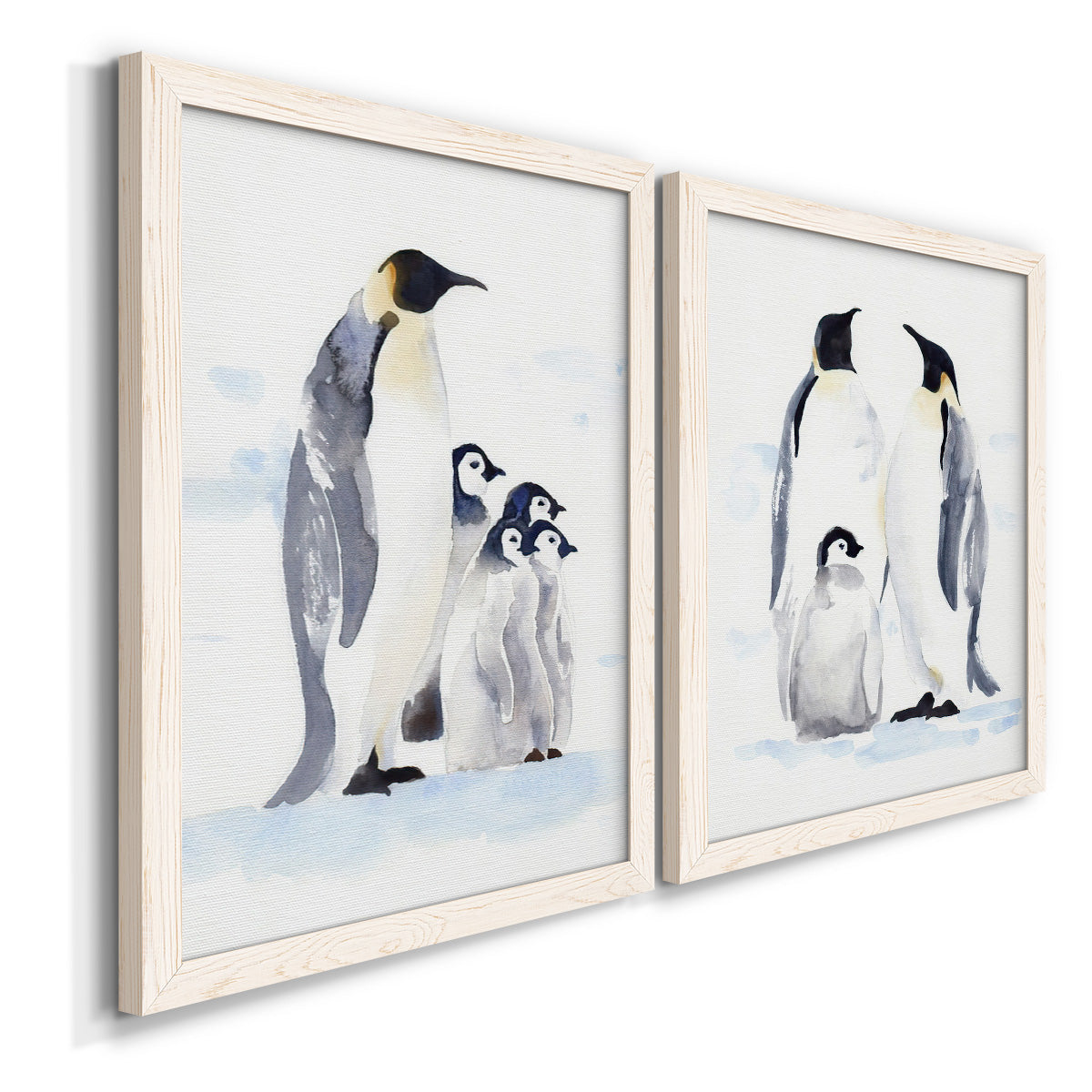 Angled view of a set of two white barnwood framed canvas prints featuring two soft watercolor wildlife illustrations featuring adult emperor penguins with fluffy chicks.