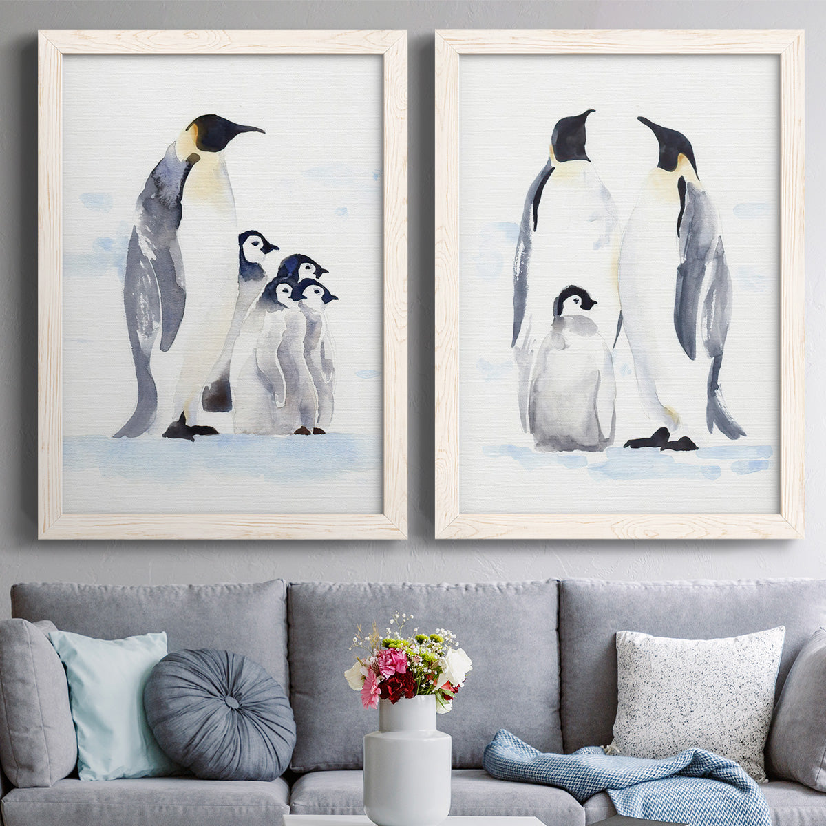 Set of two white barnwood framed canvas prints depicting paired soft watercolor wildlife illustrations with black white and yellow birds standing on pale icy ground above a sofa in a contemporary living room.