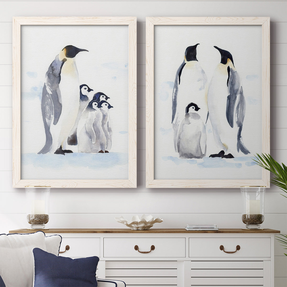 Set of two white barnwood framed canvas prints depicting a coordinated art pair of adult emperor penguins with fluffy chicks rendered as soft watercolor wildlife illustrations above a dining room sideboard.
