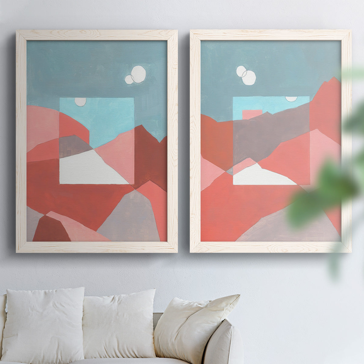 Set of two white barnwood framed canvas prints depicting faceted hills, warm horizons, and flat color planes arranged across two complementary compositions above a sofa in a bright living space.