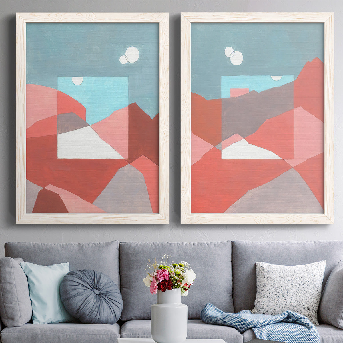 Set of two white barnwood framed canvas prints depicting paired modern abstract landscapes with faceted hills, warm horizons, and flat color planes above a sofa in a contemporary living room.