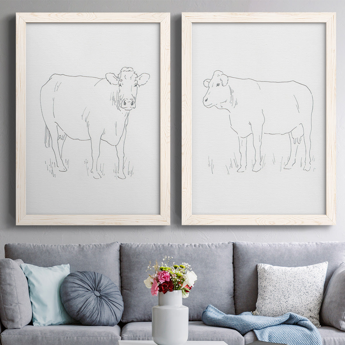 Set of two white barnwood framed canvas prints depicting paired minimalist livestock sketches with fine gray lines, white backgrounds, and quiet farm-animal profiles above a sofa in a contemporary living room.