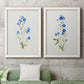 Set of two white barnwood framed canvas prints depicting a coordinated art pair of small blue wildflower stems rendered as delicate watercolor botanical studies above a sofa in a modern living room.