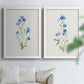 Set of two white barnwood framed canvas prints depicting tiny cobalt blossoms, fine green leaves, and abundant white space arranged across two complementary compositions above a sofa in a bright living space.