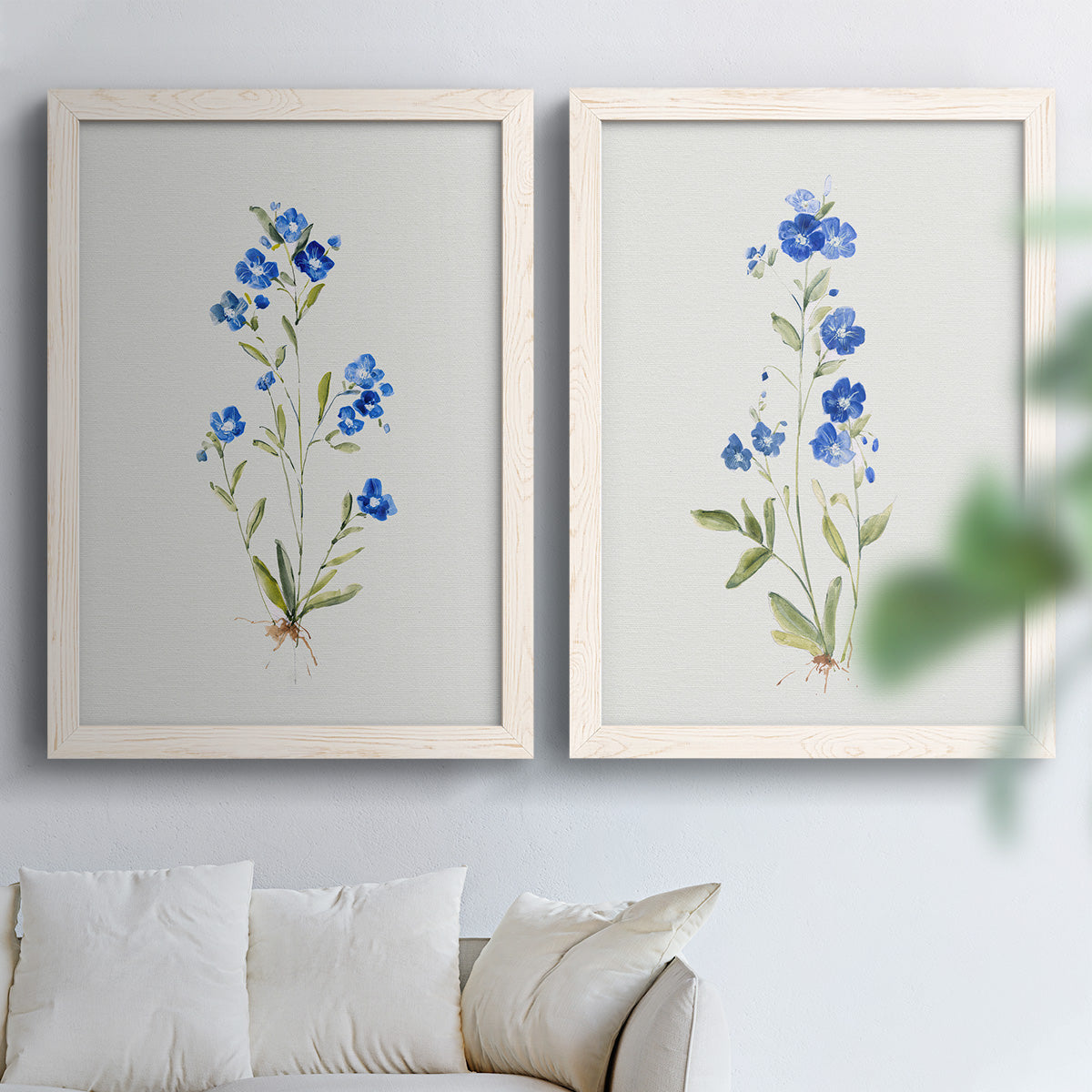Set of two white barnwood framed canvas prints depicting tiny cobalt blossoms, fine green leaves, and abundant white space arranged across two complementary compositions above a sofa in a bright living space.