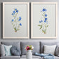 Set of two white barnwood framed canvas prints depicting paired delicate watercolor botanical studies with tiny cobalt blossoms, fine green leaves, and abundant white space above a sofa in a contemporary living room.
