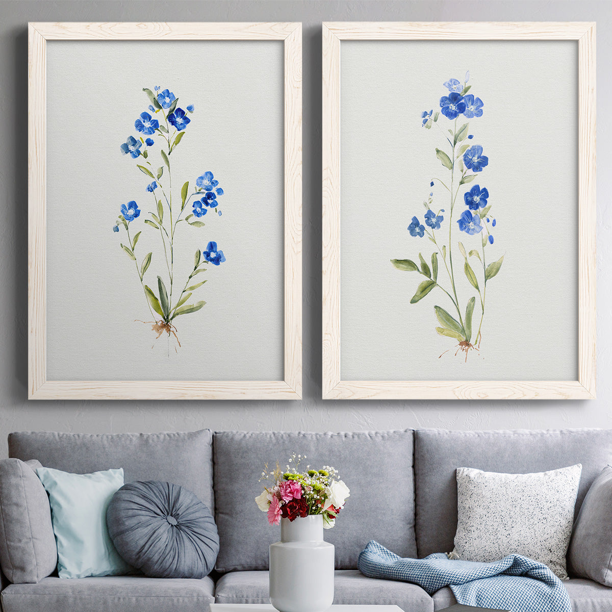 Set of two white barnwood framed canvas prints depicting paired delicate watercolor botanical studies with tiny cobalt blossoms, fine green leaves, and abundant white space above a sofa in a contemporary living room.