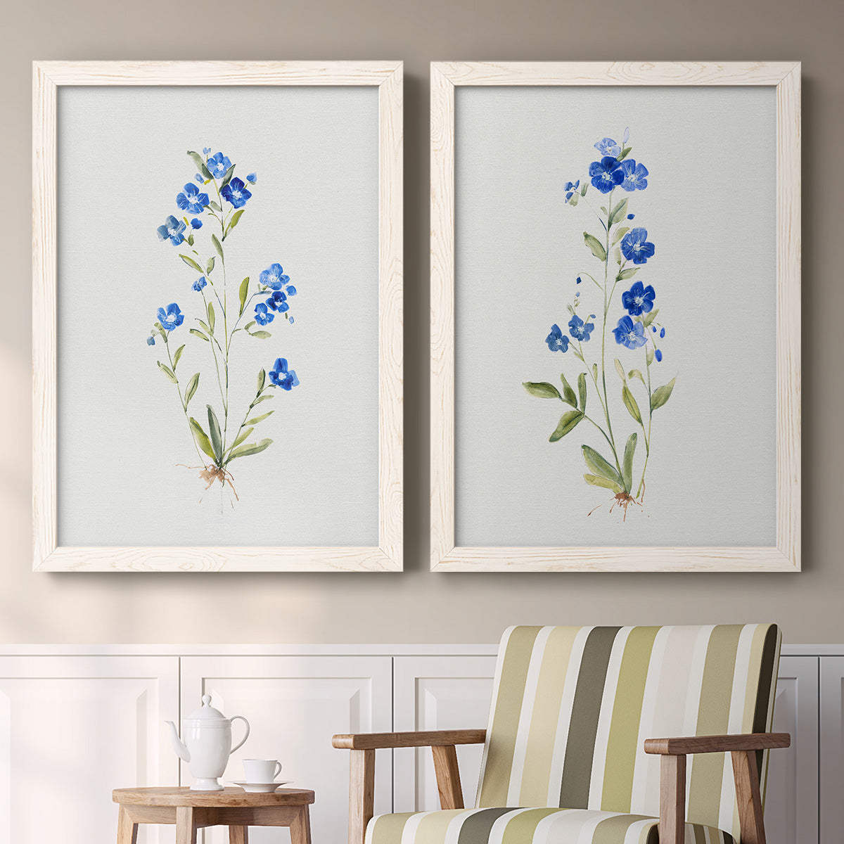 Set of two white barnwood framed canvas prints depicting two delicate watercolor botanical studies featuring small blue wildflower stems above a sideboard in a cozy sitting room.