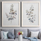 Set of two white barnwood framed canvas prints depicting paired minimalist watercolor botanical studies with soft sage gray and muted green leaves on clean white grounds above a sofa in a contemporary living room.