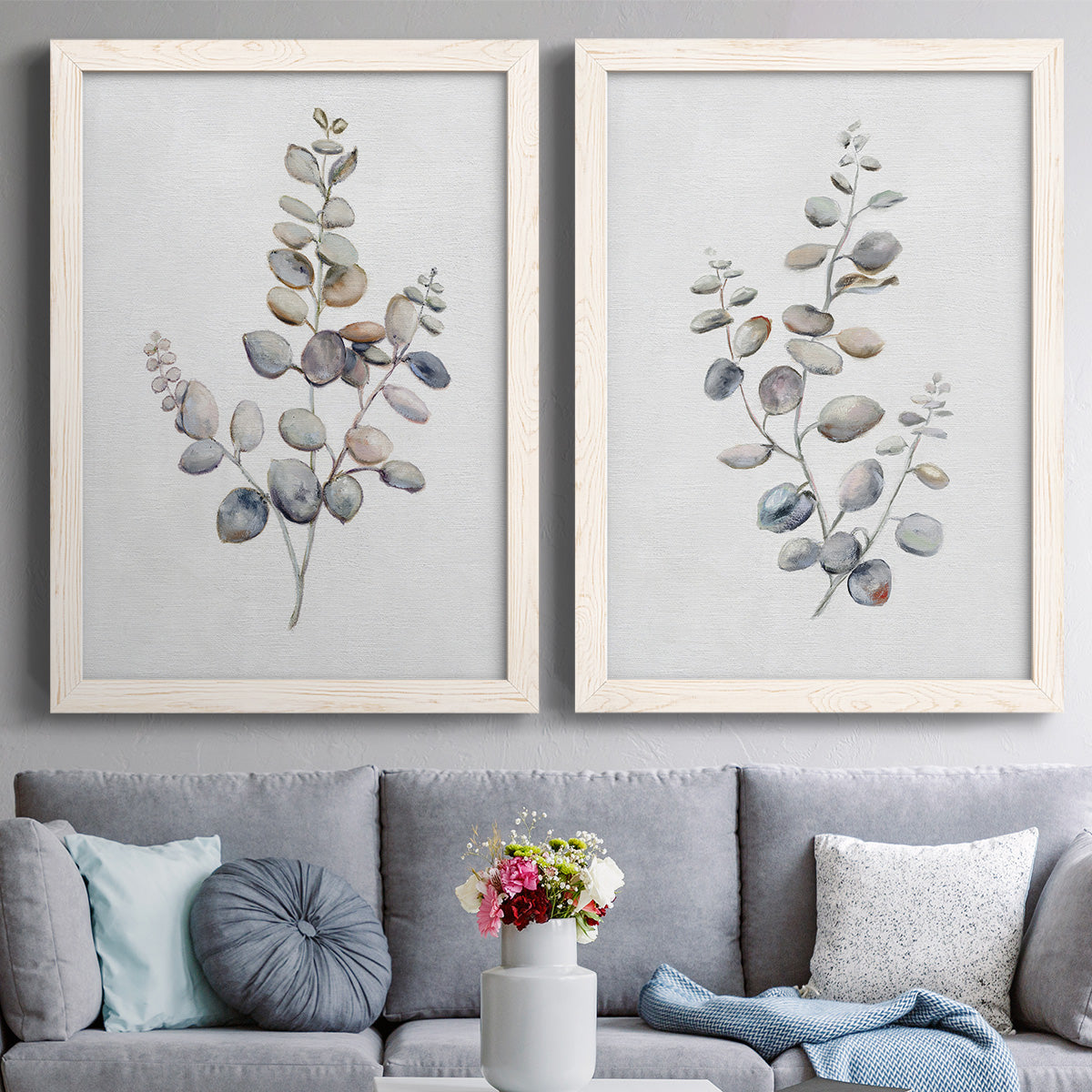 Set of two white barnwood framed canvas prints depicting paired minimalist watercolor botanical studies with soft sage gray and muted green leaves on clean white grounds above a sofa in a contemporary living room.