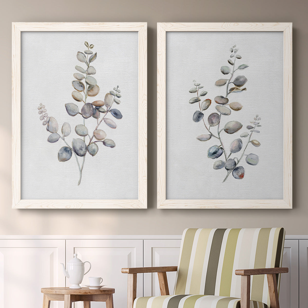 Set of two white barnwood framed canvas prints depicting two minimalist watercolor botanical studies featuring delicate eucalyptus branches with rounded leaves above a sideboard in a cozy sitting room.