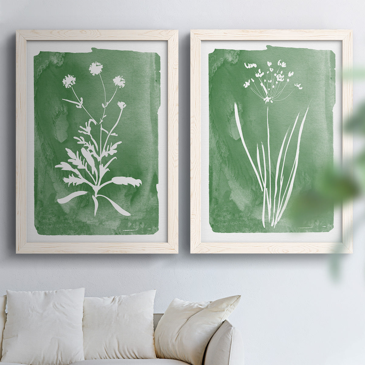 Set of two white barnwood framed canvas prints depicting fine green branches, sparse blue or white blossoms, and cream grounds arranged across two complementary compositions above a sofa in a bright living space.