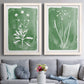 Set of two white barnwood framed canvas prints depicting paired rustic minimalist botanical studies with fine green branches, sparse blue or white blossoms, and cream grounds above a sofa in a contemporary living room.