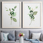 Set of two white barnwood framed canvas prints depicting paired rustic watercolor botanical studies with dark round berries, delicate stems, and clean white space above a sofa in a contemporary living room.
