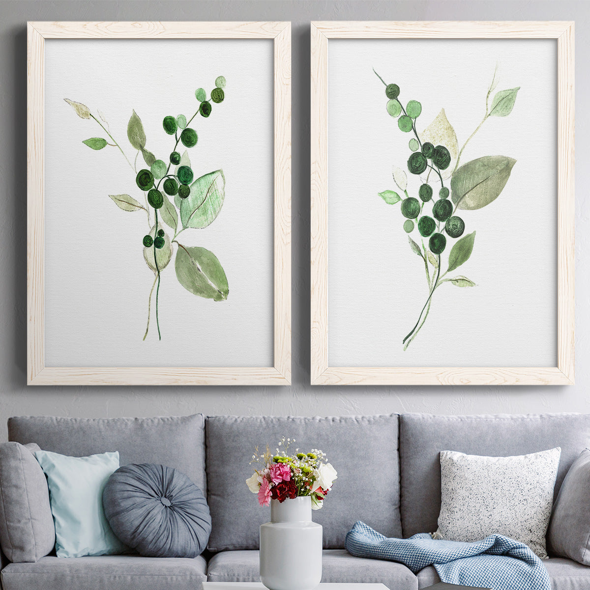 Set of two white barnwood framed canvas prints depicting paired rustic watercolor botanical studies with dark round berries, delicate stems, and clean white space above a sofa in a contemporary living room.