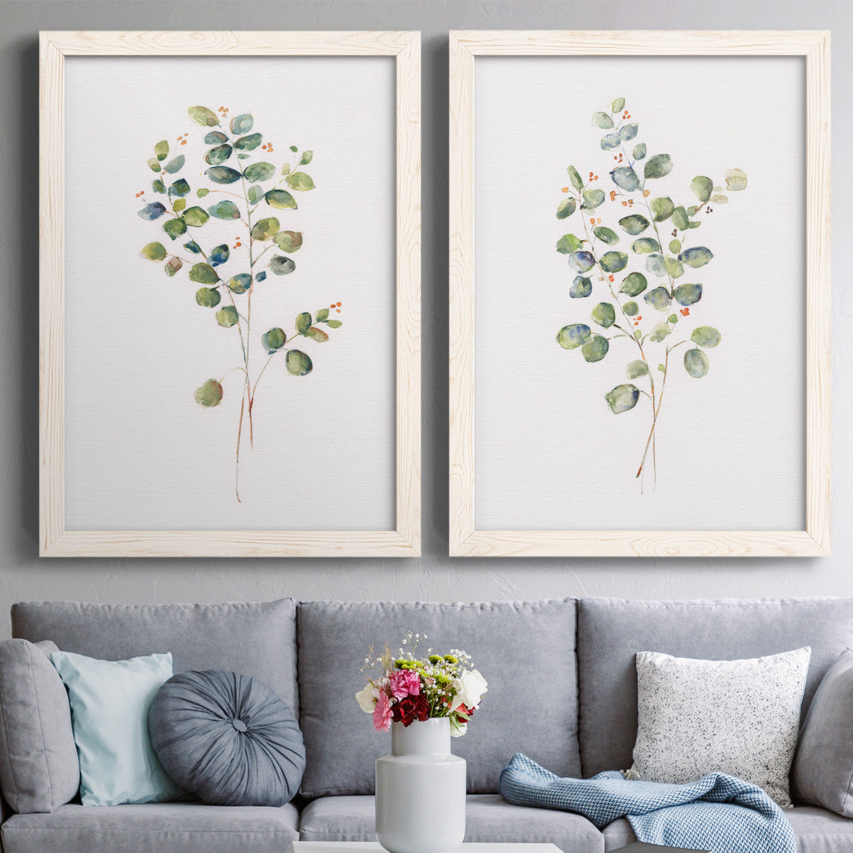 Set of two white barnwood framed canvas prints depicting paired minimalist watercolor botanical studies with soft sage gray and muted green leaves on clean white grounds above a sofa in a contemporary living room.