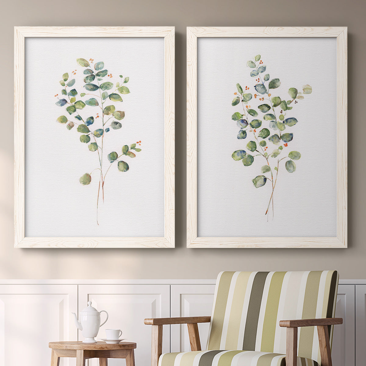 Set of two white barnwood framed canvas prints depicting two minimalist watercolor botanical studies featuring delicate eucalyptus branches with rounded leaves above a sideboard in a cozy sitting room.