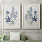 Set of two white barnwood framed canvas prints depicting a coordinated art pair of blue fern fronds and small woodland flowers rendered as rustic botanical illustrations above a sofa in a modern living room.