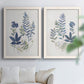 Set of two white barnwood framed canvas prints depicting layered leaves, indigo sprigs, and lightly textured cream grounds arranged across two complementary compositions above a sofa in a bright living space.