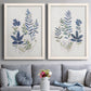 Set of two white barnwood framed canvas prints depicting paired rustic botanical illustrations with layered leaves, indigo sprigs, and lightly textured cream grounds above a sofa in a contemporary living room.