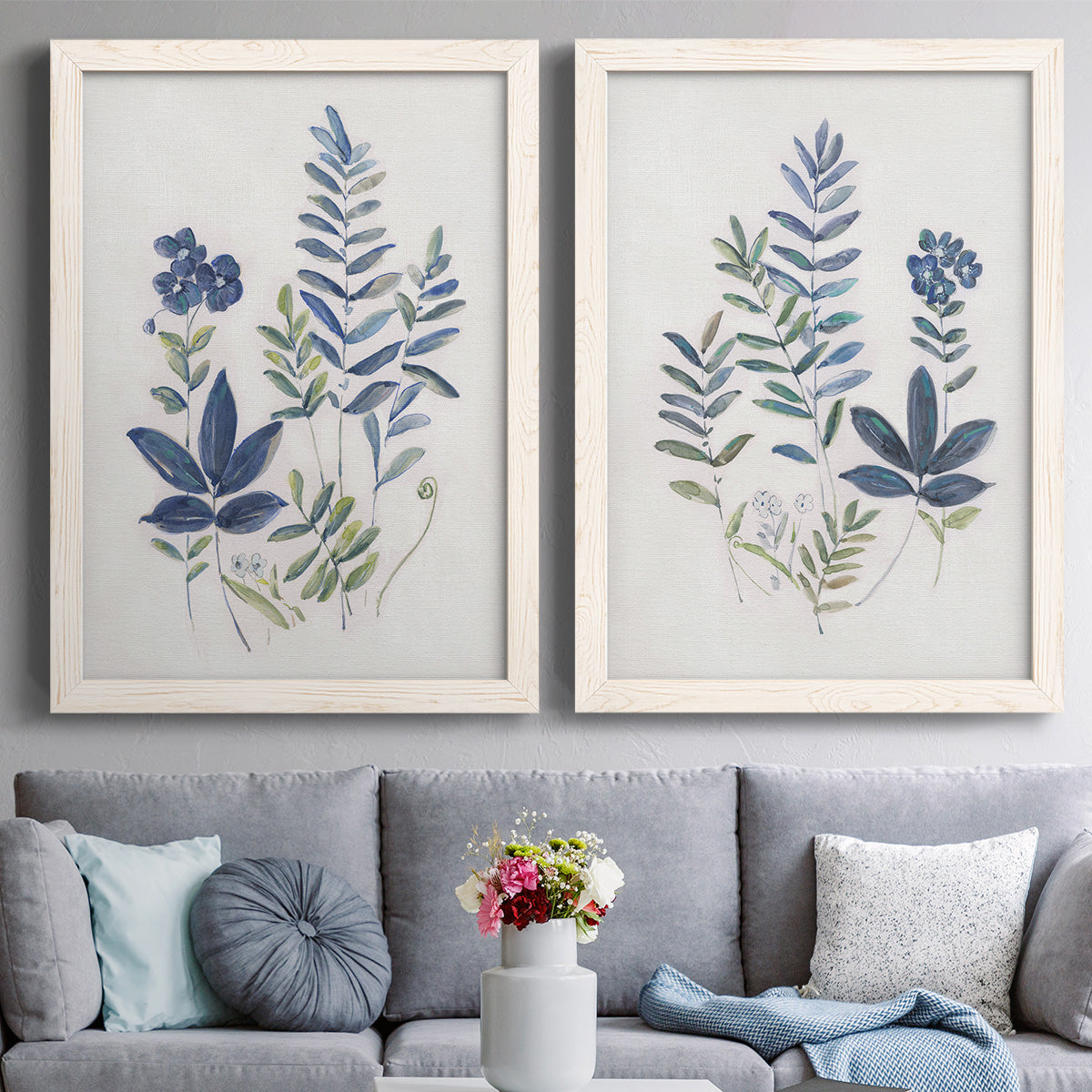 Set of two white barnwood framed canvas prints depicting paired rustic botanical illustrations with layered leaves, indigo sprigs, and lightly textured cream grounds above a sofa in a contemporary living room.