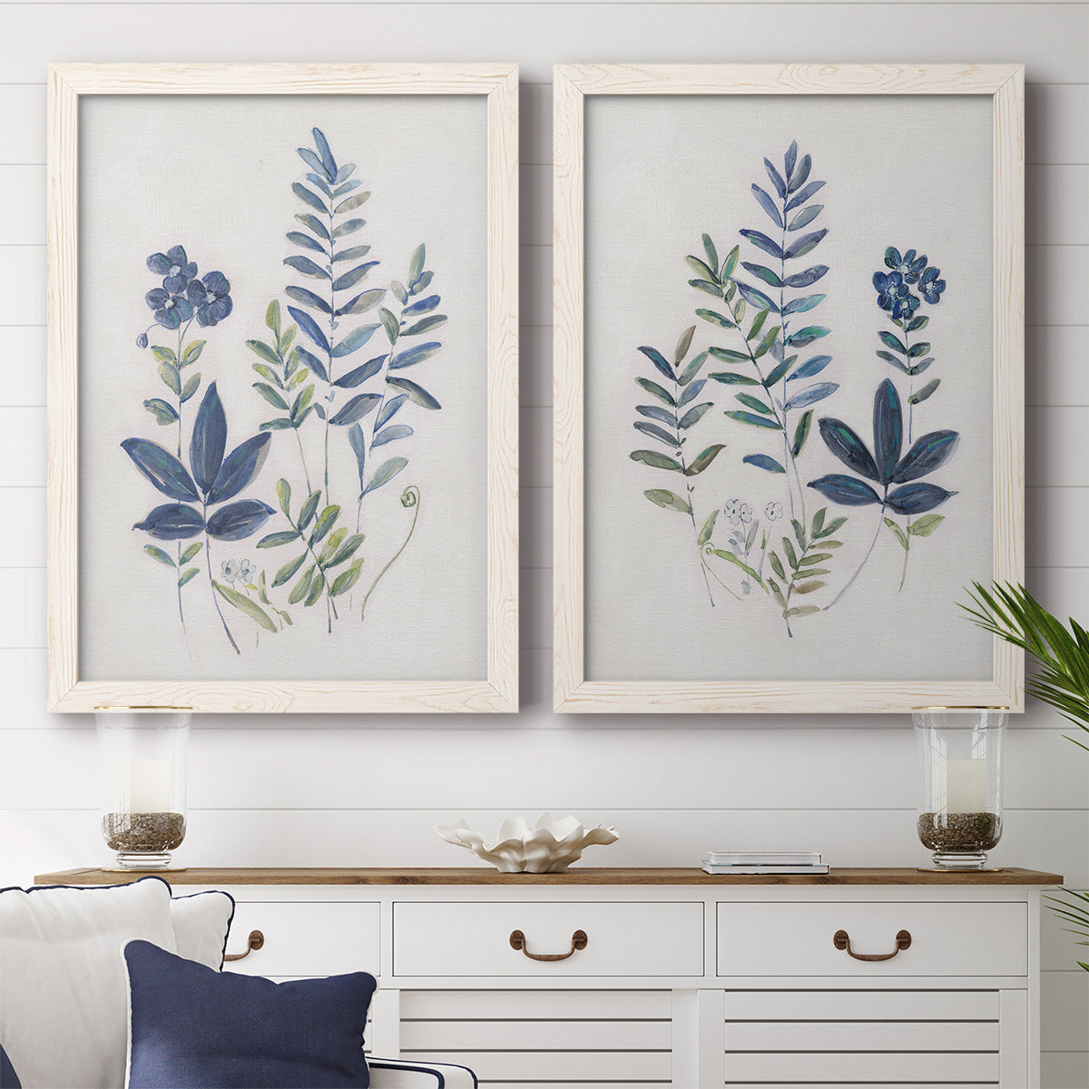 Set of two white barnwood framed canvas prints depicting a coordinated art pair of blue fern fronds and small woodland flowers rendered as rustic botanical illustrations above a dining room sideboard.