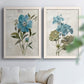 Set of two white barnwood framed canvas prints depicting large blue flowers, muted green leaves, and linen-textured backgrounds arranged across two complementary compositions above a sofa in a bright living space.