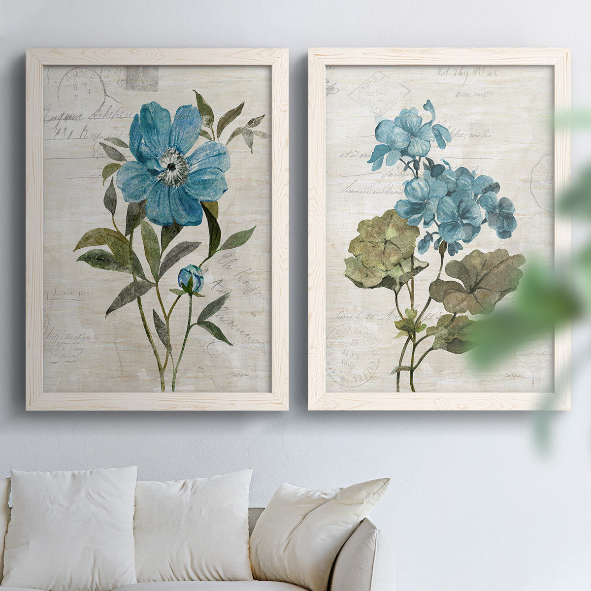 Set of two white barnwood framed canvas prints depicting large blue flowers, muted green leaves, and linen-textured backgrounds arranged across two complementary compositions above a sofa in a bright living space.