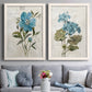 Set of two white barnwood framed canvas prints depicting paired rustic floral illustrations with large blue flowers, muted green leaves, and linen-textured backgrounds above a sofa in a contemporary living room.