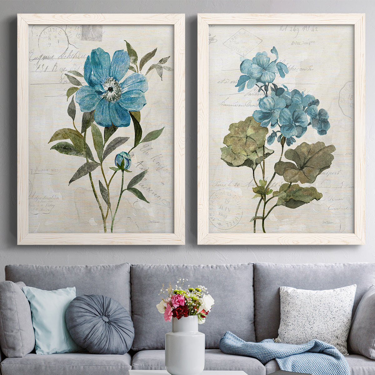 Set of two white barnwood framed canvas prints depicting paired rustic floral illustrations with large blue flowers, muted green leaves, and linen-textured backgrounds above a sofa in a contemporary living room.