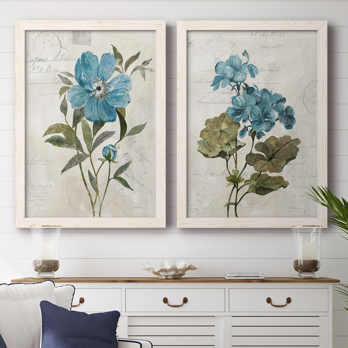 Set of two white barnwood framed canvas prints depicting a coordinated art pair of blue peony and hydrangea-like blooms over vintage script rendered as rustic floral illustrations above a dining room sideboard.