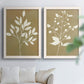 Set of two white barnwood framed canvas prints depicting simple white botanical forms featuring broad leaves and delicate buds on golden brown backgrounds above a sofa in a bright living space.