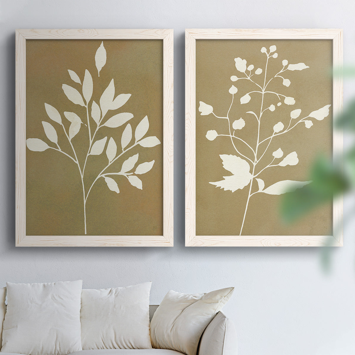 Set of two white barnwood framed canvas prints depicting simple white botanical forms featuring broad leaves and delicate buds on golden brown backgrounds above a sofa in a bright living space.