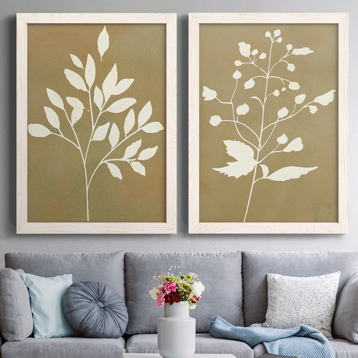 Set of two white barnwood framed canvas prints depicting coordinated foliage silhouettes in ivory tan and antique gold with clean modern spacing above a sofa in a contemporary living room.
