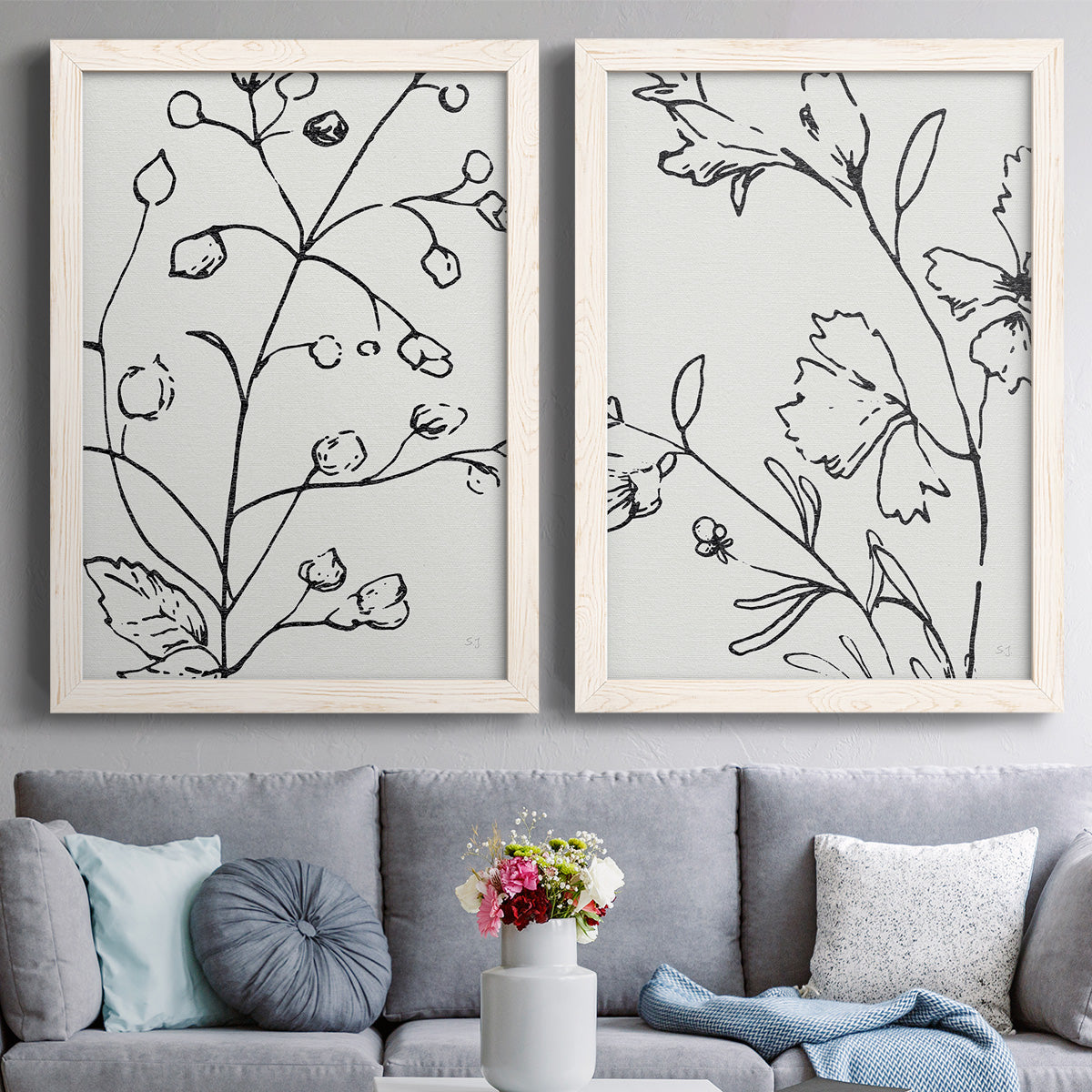 Set of two white barnwood framed canvas prints depicting paired minimalist botanical line art with fine monochrome outlines, cropped stems, and clean white fields above a sofa in a contemporary living room.