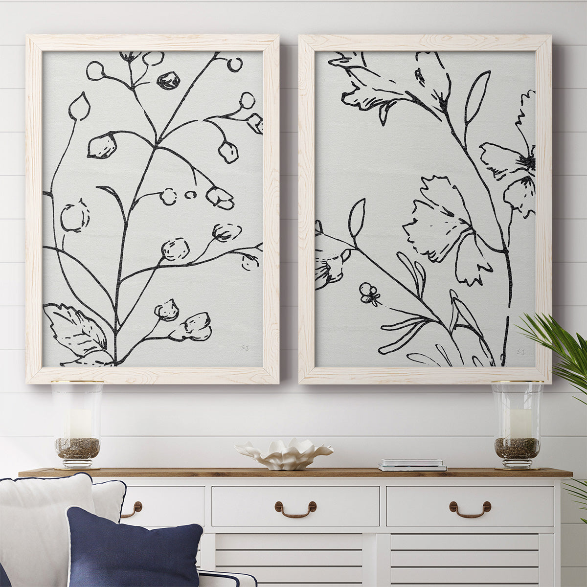 Set of two white barnwood framed canvas prints depicting a coordinated art pair of black contour drawings of leaves and flowers rendered as minimalist botanical line art above a dining room sideboard.