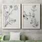 Set of two white barnwood framed canvas prints depicting a coordinated art pair of black line drawings of wildflowers and leaves rendered as minimalist botanical sketches above a sofa in a modern living room.