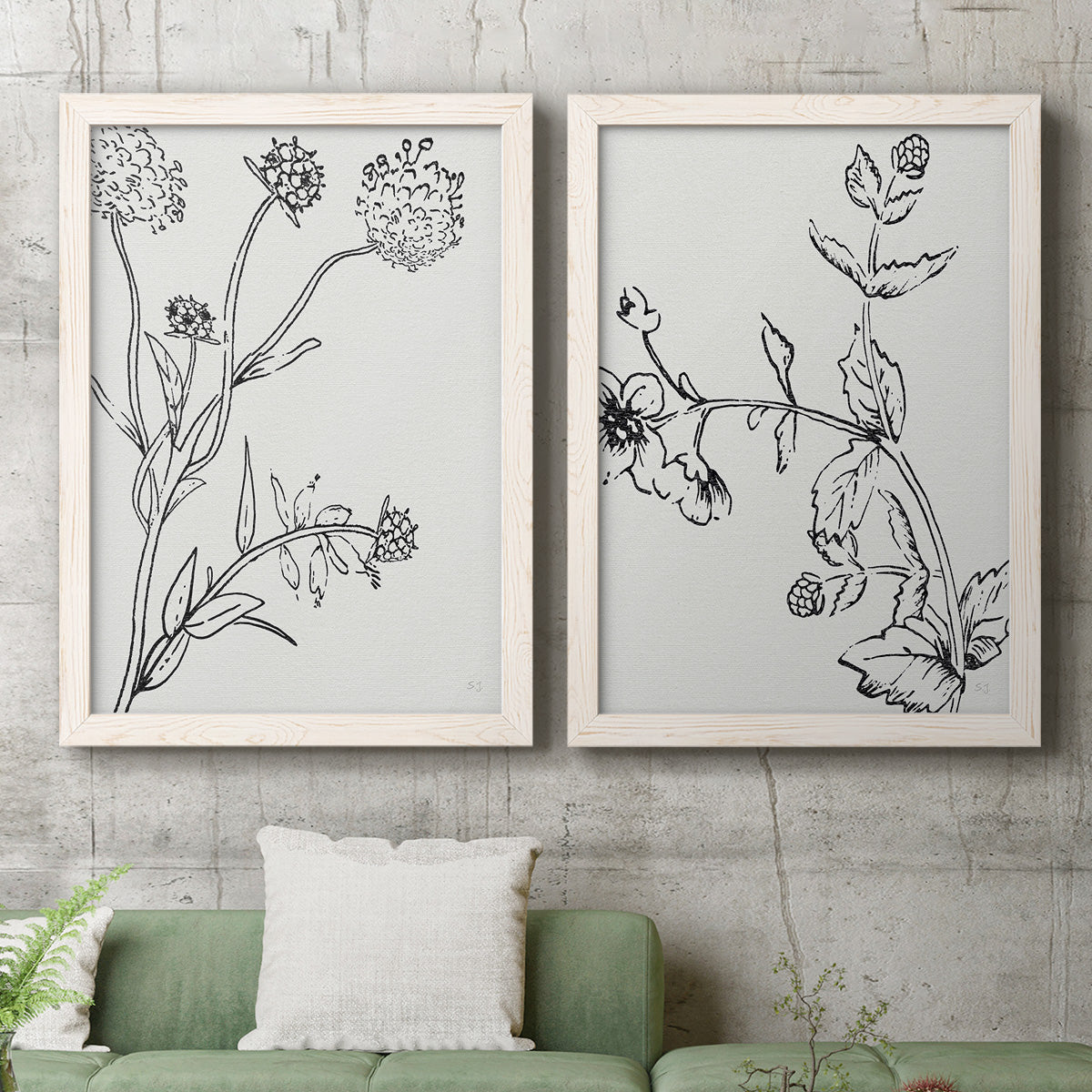 Set of two white barnwood framed canvas prints depicting a coordinated art pair of black line drawings of wildflowers and leaves rendered as minimalist botanical sketches above a sofa in a modern living room.