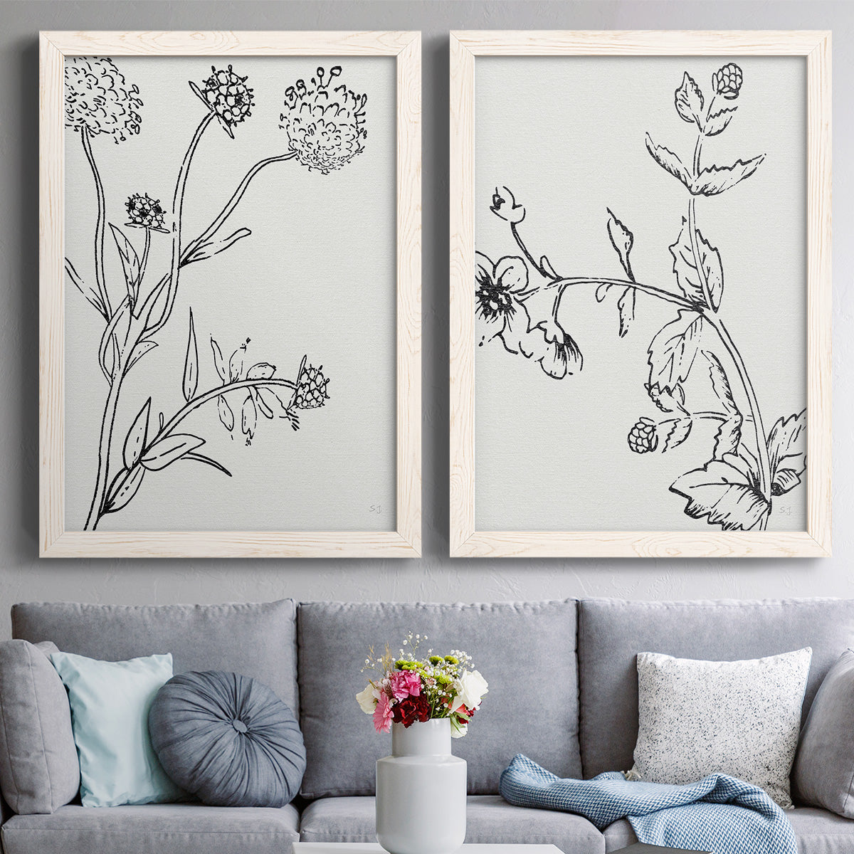 Set of two white barnwood framed canvas prints depicting paired minimalist botanical sketches with fine stems, seed heads, blossoms, and open white backgrounds above a sofa in a contemporary living room.