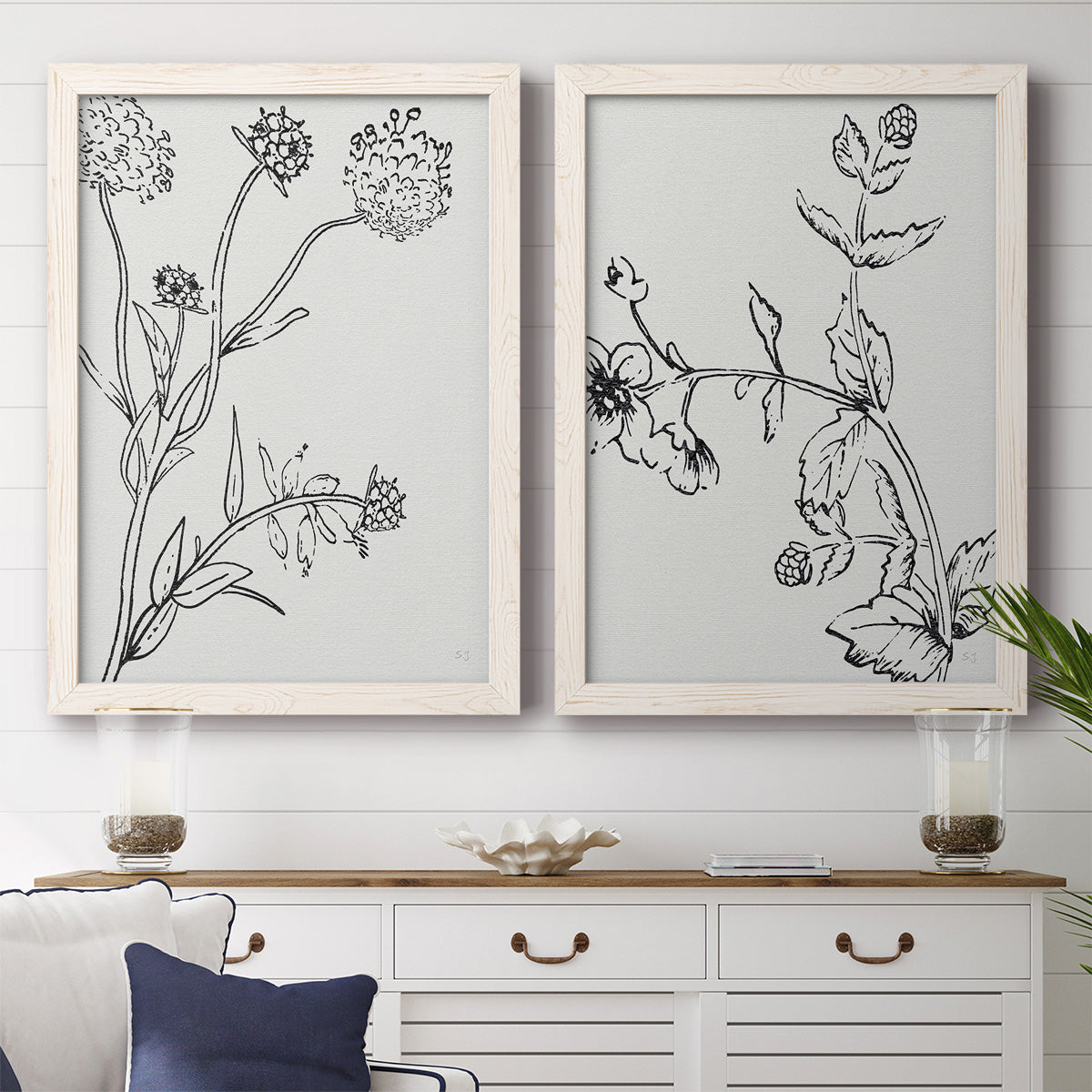 Set of two white barnwood framed canvas prints depicting a coordinated art pair of black line drawings of wildflowers and leaves rendered as minimalist botanical sketches above a dining room sideboard.
