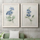 Set of two white barnwood framed canvas prints depicting a coordinated art pair of blue periwinkle flowers with leafy stems rendered as farmhouse botanical illustrations above a sofa in a modern living room.