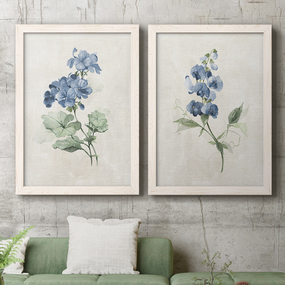 Set of two white barnwood framed canvas prints depicting a coordinated art pair of blue periwinkle flowers with leafy stems rendered as farmhouse botanical illustrations above a sofa in a modern living room.