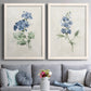 Set of two white barnwood framed canvas prints depicting paired farmhouse botanical illustrations with single blue blossoms, green leaves, and lightly textured cream grounds above a sofa in a contemporary living room.