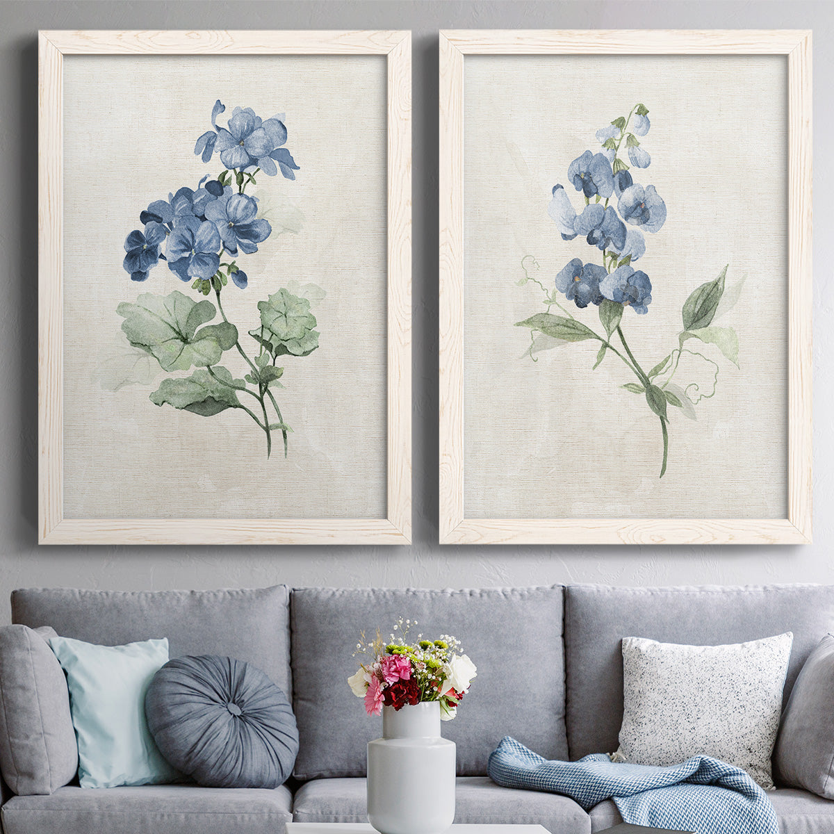 Set of two white barnwood framed canvas prints depicting paired farmhouse botanical illustrations with single blue blossoms, green leaves, and lightly textured cream grounds above a sofa in a contemporary living room.