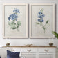 Set of two white barnwood framed canvas prints depicting a coordinated art pair of blue periwinkle flowers with leafy stems rendered as farmhouse botanical illustrations above a dining room sideboard.