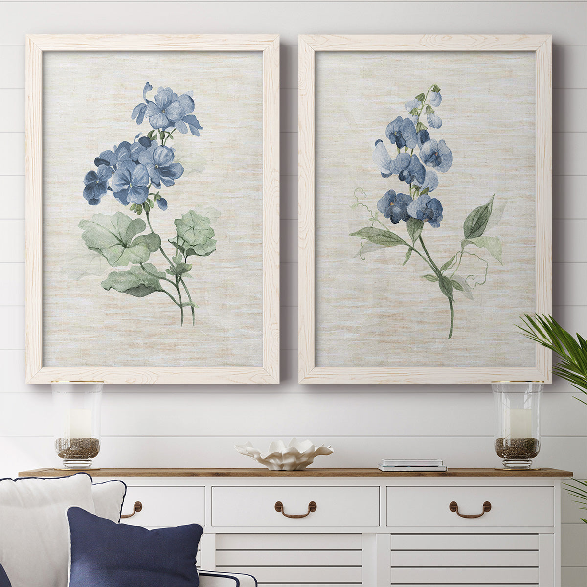 Set of two white barnwood framed canvas prints depicting a coordinated art pair of blue periwinkle flowers with leafy stems rendered as farmhouse botanical illustrations above a dining room sideboard.
