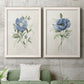 Set of two white barnwood framed canvas prints depicting a coordinated art pair of blue periwinkle flowers with leafy stems rendered as farmhouse botanical illustrations above a sofa in a modern living room.
