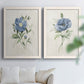 Set of two white barnwood framed canvas prints depicting single blue blossoms, green leaves, and lightly textured cream grounds arranged across two complementary compositions above a sofa in a bright living space.