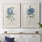 Set of two white barnwood framed canvas prints depicting a coordinated art pair of blue periwinkle flowers with leafy stems rendered as farmhouse botanical illustrations above a dining room sideboard.
