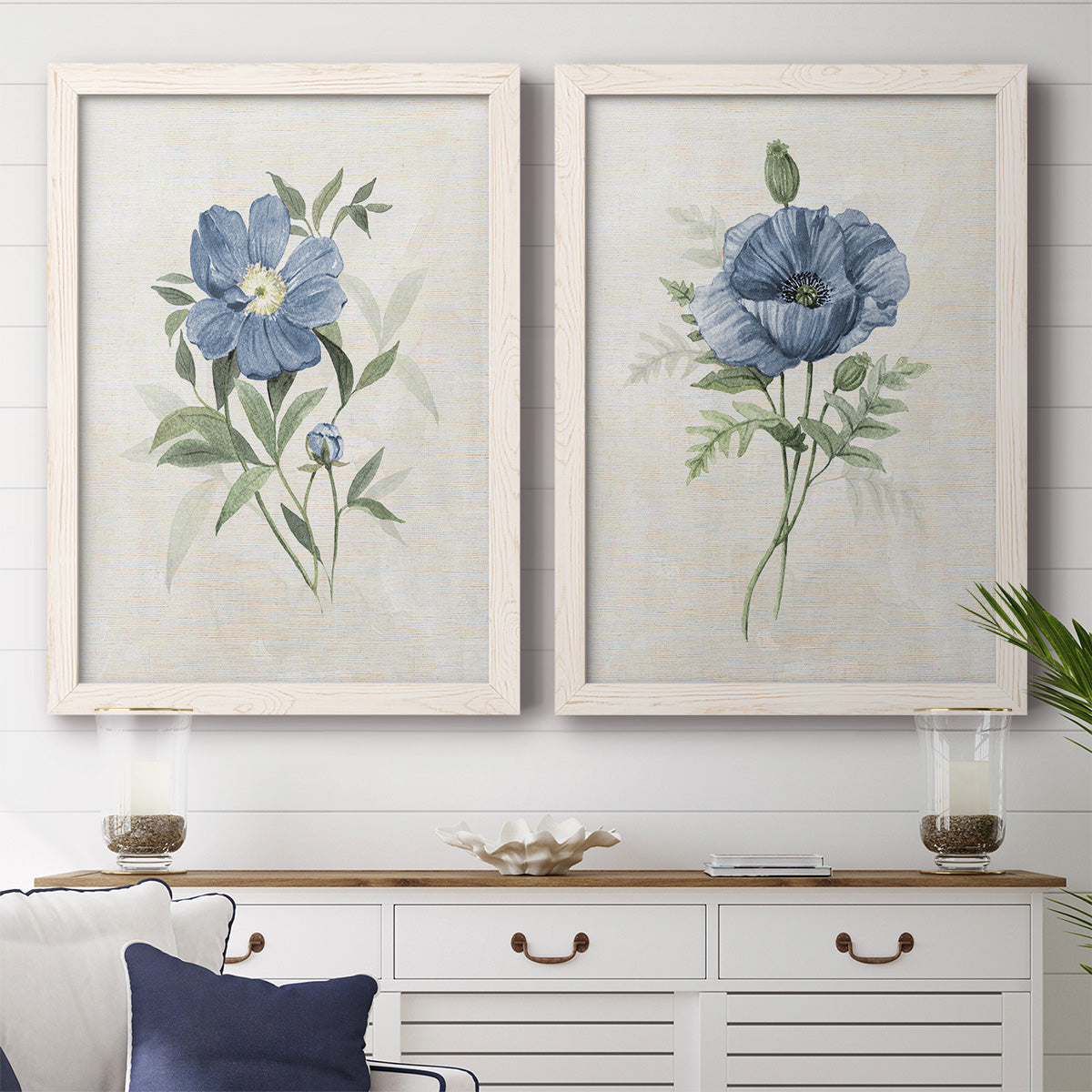 Set of two white barnwood framed canvas prints depicting a coordinated art pair of blue periwinkle flowers with leafy stems rendered as farmhouse botanical illustrations above a dining room sideboard.